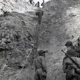 U. S. Army Rangers show how they accomplished their feat by climbing a makeshift ladder to take German gun crews by surprise.