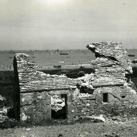 Through the battered roof of a building on the French invasion coast can be seen ships of the Allies amphibious fleet.