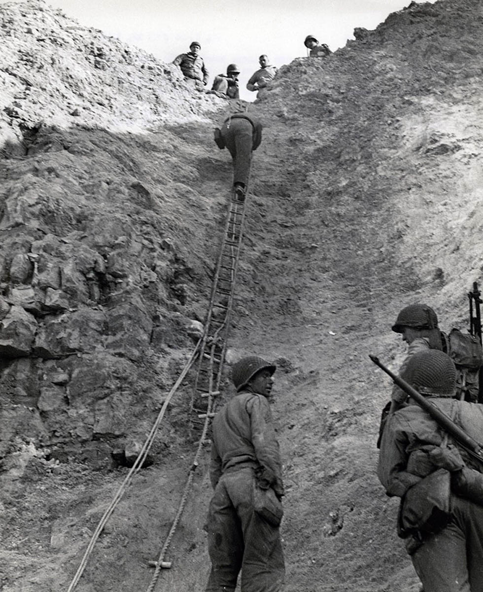 U. S. Army Rangers show how they accomplished their feat by climbing a makeshift ladder to take German gun crews by surprise.