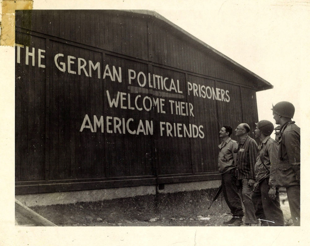 Survivor and American troops look at sign on building, welcoming American liberators.