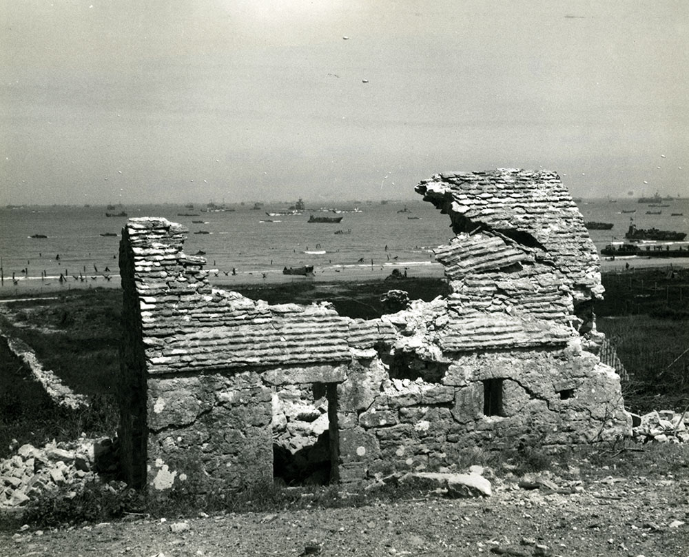 Through the battered roof of a building on the French invasion coast can be seen ships of the Allies amphibious fleet.