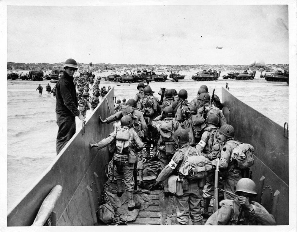 The photograph shows soldiers exiting an amphibious landing craft into shallow water near a beach. Tanks and other vehicles can be seen in the background on the beach.