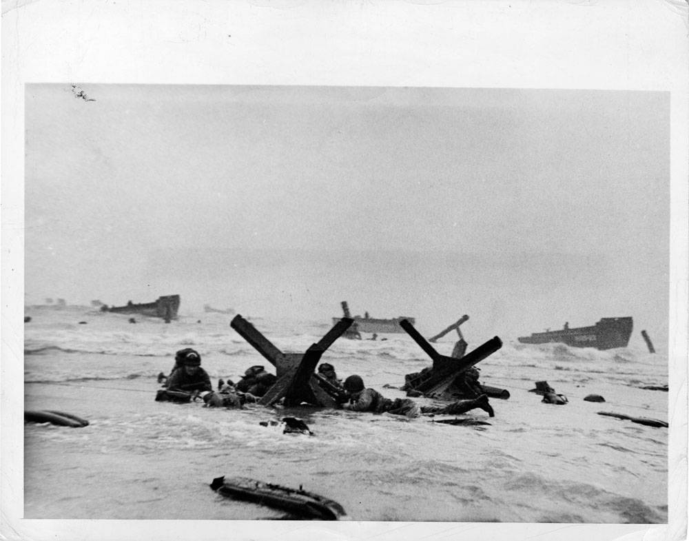 Steel and concrete obstacles, put up by the Germans to trap invasion craft, didn't stop Allied assault troops going ashore during the invasion of the French coast. To the contrary, these obstacles served as cover against enemy fire. Here, U.S. and British troops huddle against one of the "traps" as protection against enemy machine gun fire.