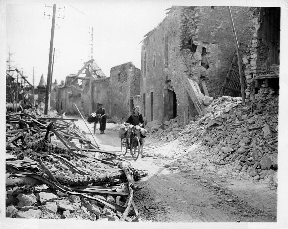 A Frenchmen and his wife, loaded milk cans on the handlebars of their bicycles, make their way through the wreckage of this important city in Normandy.