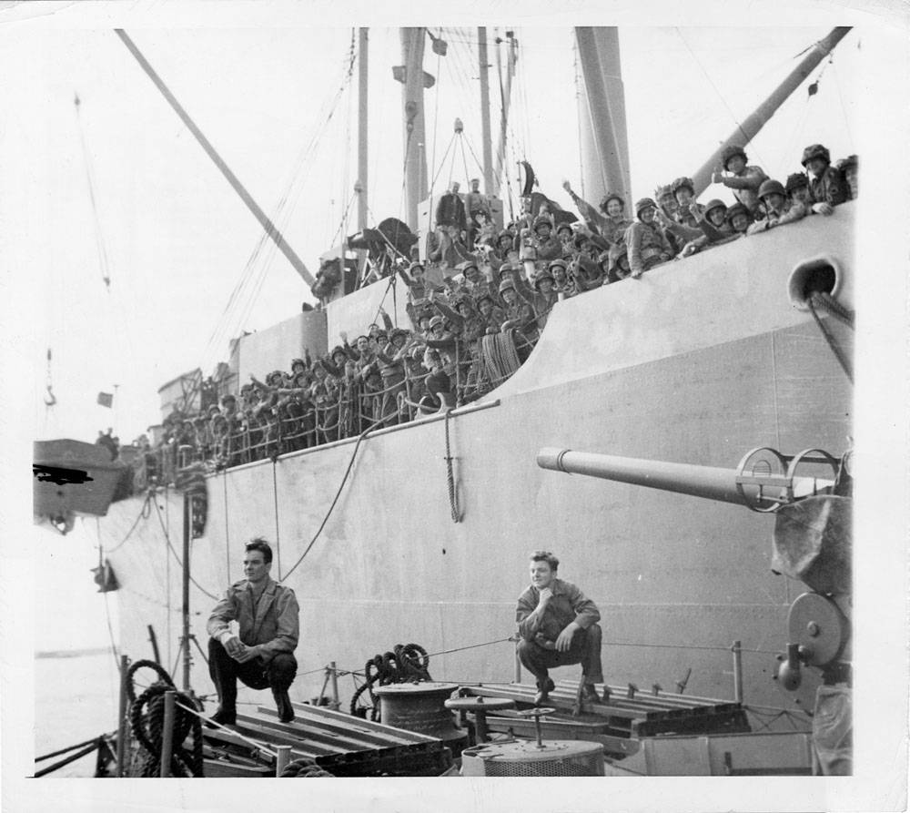 Allied assault troops wave jubilantly from aboard a transport which is pulling out from a British embarkation point enroute to France and the long-awaited invasion.
