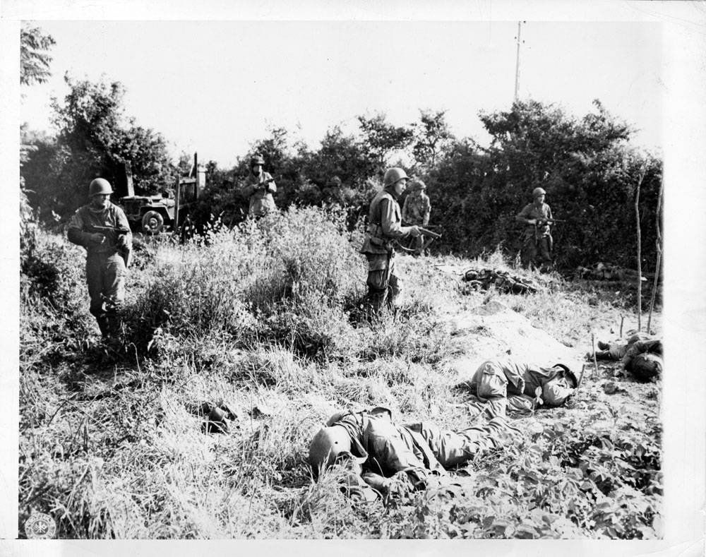 Moving warily through a field, American paratroopers pass members of their own outfit who were victims of snipers at Carentan. Their faces lined with determination, the sky-fighters push ahead, seeking revenge.