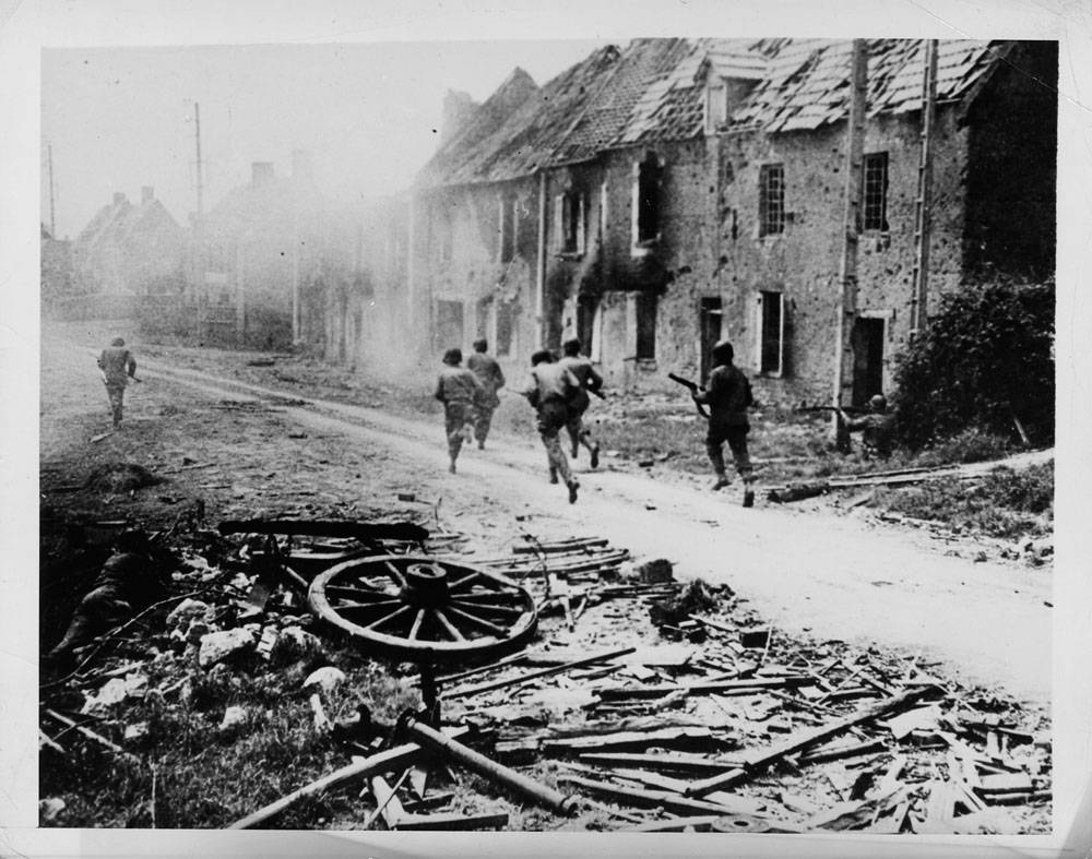 Under the cover of comrades' fire at left and near the building at right, a group of American soldiers dash through the haze of battle smoke to attack hidden Nazis in a small French village.