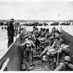 The photograph shows soldiers exiting an amphibious landing craft into shallow water near a beach. Tanks and other vehicles can be seen in the background on the beach.