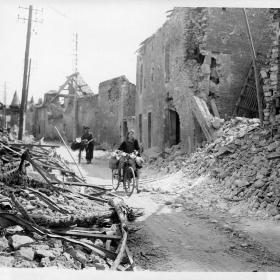 A Frenchmen and his wife, loaded milk cans on the handlebars of their bicycles, make their way through the wreckage of this important city in Normandy.