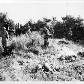 Moving warily through a field, American paratroopers pass members of their own outfit who were victims of snipers at Carentan. Their faces lined with determination, the sky-fighters push ahead, seeking revenge.