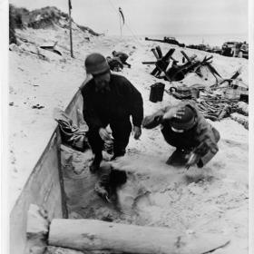 Moving 'on the double' these members of a U.S. Navy beach battaluon [sic] dive for the protection offered by a ditch as a Nazi plane swoops down to strafe the shore somewhere in Normandy.