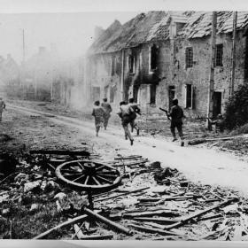 Under the cover of comrades' fire at left and near the building at right, a group of American soldiers dash through the haze of battle smoke to attack hidden Nazis in a small French village.